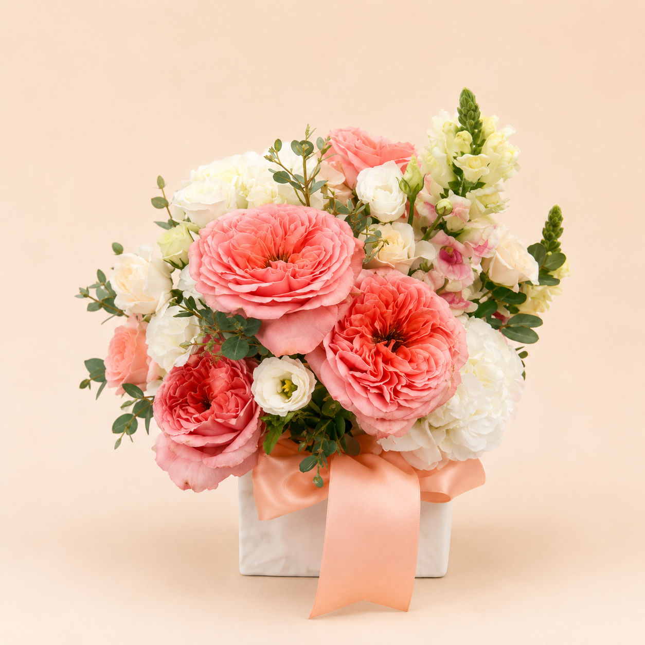 A floral arrangement in a white ceramic vase a peach ribbon, featuring large pink and coral roses, white flowers, and green foliage on a soft beige background.