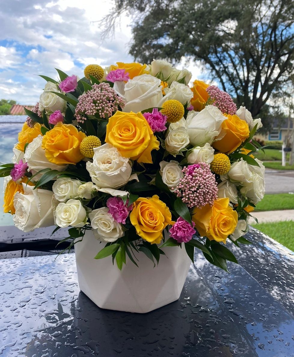 Colorful flower arrangement with yellow, white, and pink flowers in a geometric vase on a car hood.