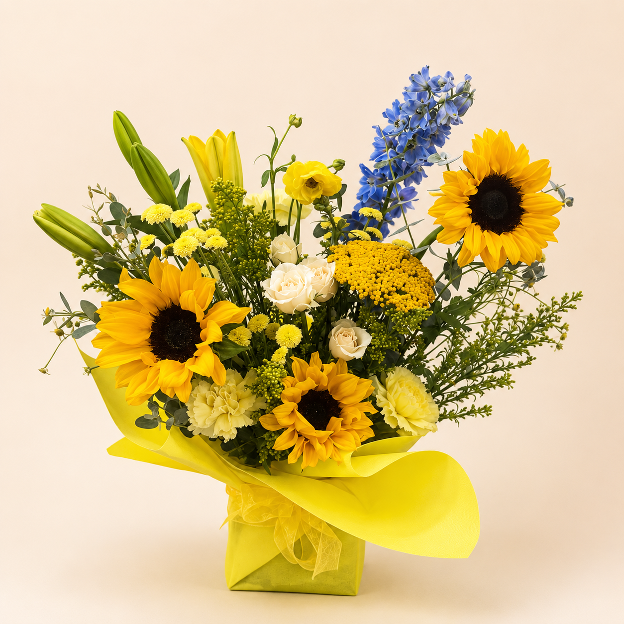 A vibrant floral bouquet featuring yellow sunflowers, blue delphiniums, white roses, and green foliage, wrapped in bright yellow tissue paper and tied with a matching ribbon, set against a solid light beige background.