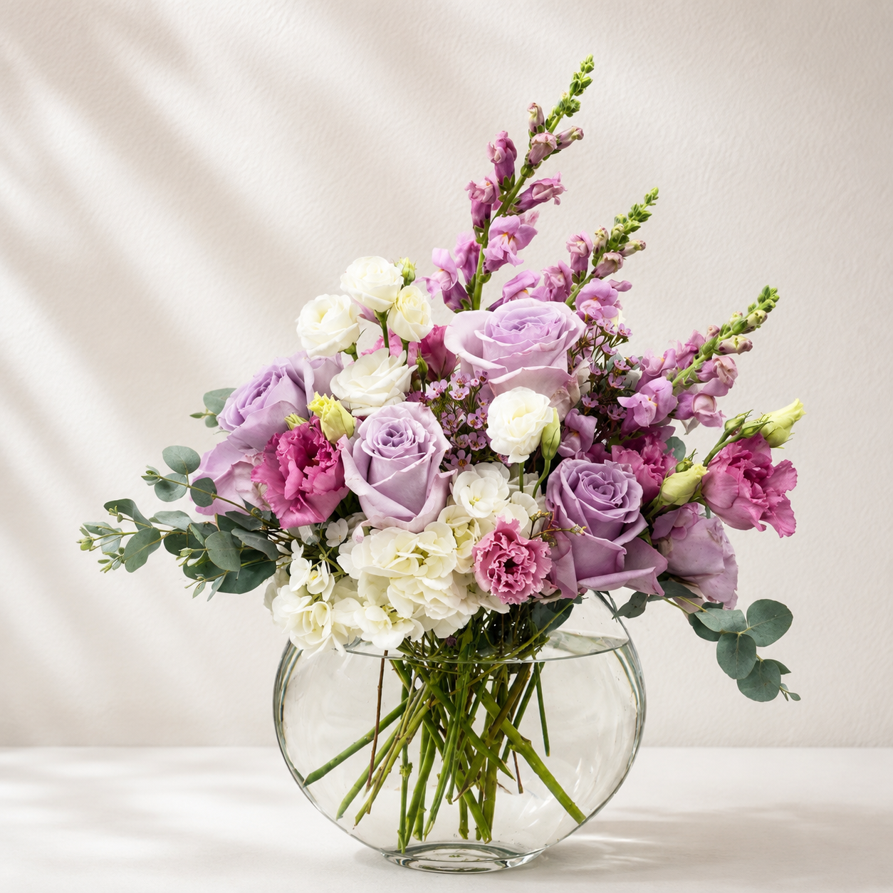 A vibrant artificial flower bouquet in a clear glass vase, featuring light purple and magenta roses, white carnations, pink carnations, and green eucalyptus stems, set against a neutral beige background with soft shadow overlays.