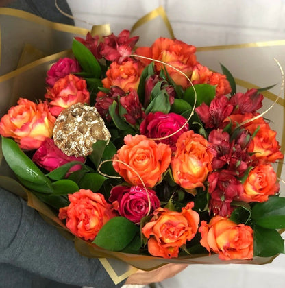Bouquet of orange and pink roses with a gold leaf in a white container.