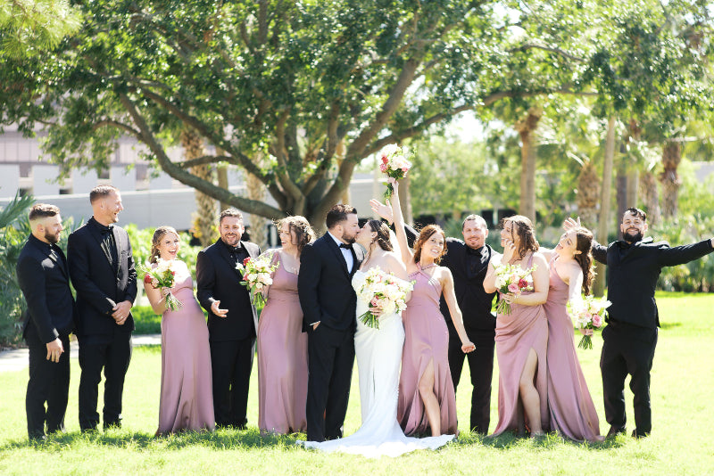 Wedding party posing outdoors with greenery and a building in the background