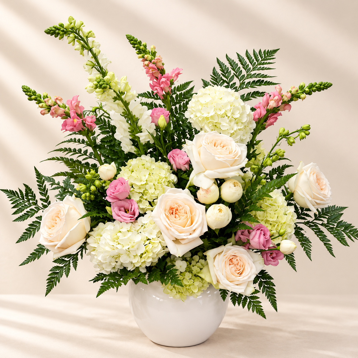 A lush floral arrangement in a vase, featuring  roses, white hydrangeas, pink snapdragons, and green foliage, set against a soft beige draped background.