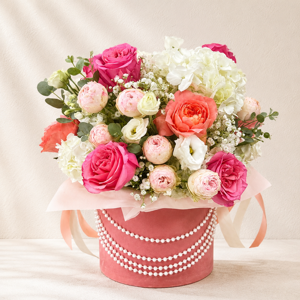 A floral arrangement of pink, coral, and white roses with green foliage, presented in a round dusty pink box decorated with white faux pearls and ribbon ties, set against a soft beige fabric backdrop.