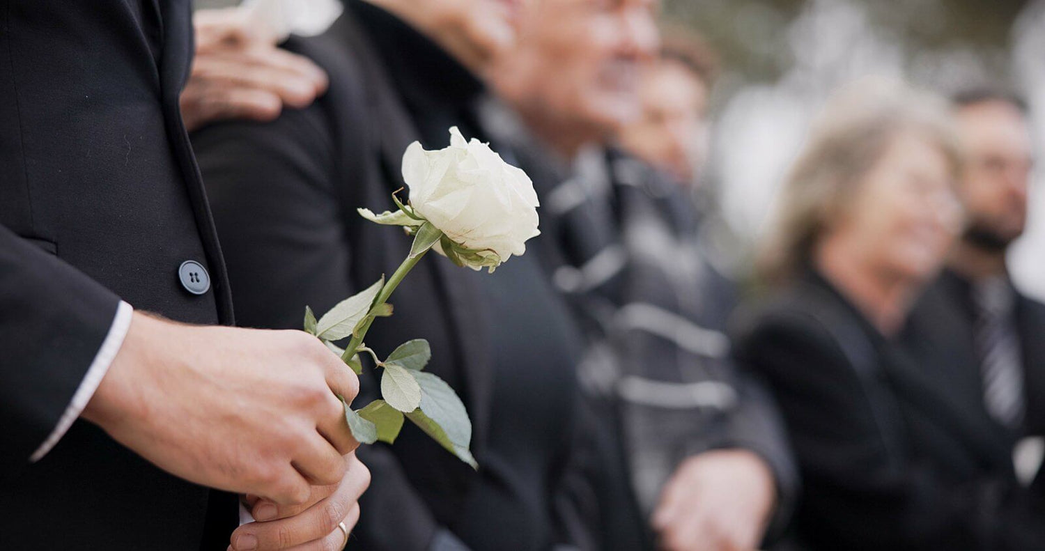 Person holding a white rose in front of a group of people, possibly at a funeral.