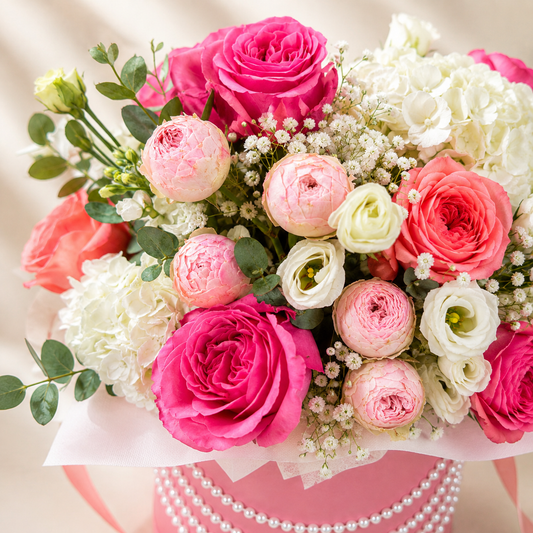 A close-up of a floral bouquet in a pink box, featuring bright pink and coral roses, blush peonies, white hydrangeas, and white baby's breath with green foliage, wrapped in pale pink paper with a pearl-like decorative band at the base.