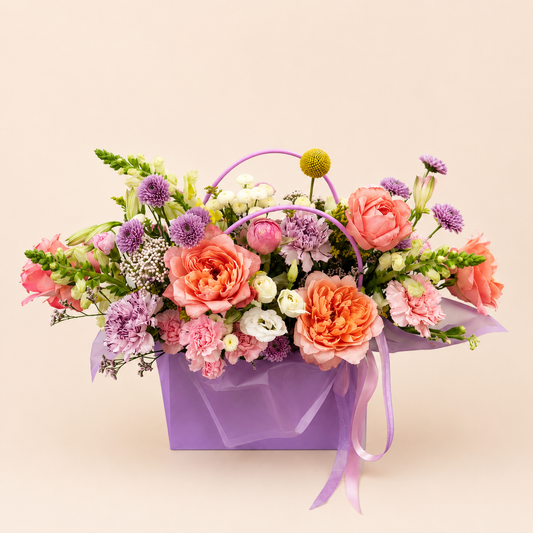 A vibrant floral arrangement in a purple gift box with a purple handle and ribbon, featuring pink and peach roses, purple chamomile, and green foliage, set against a solid light beige background.